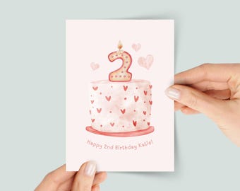 May include: A birthday card featuring a watercolor illustration of a cake with a number 2 candle and hearts. The card reads "Happy 2nd Birthday Katie!" in a soft, pink color scheme. The card is held by two hands.
