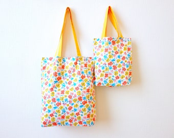 May include: Two tote bags with yellow handles and a floral print in shades of pink, blue, and yellow. The larger bag is hanging on the left and the smaller bag is hanging on the right.