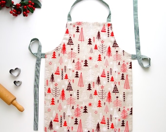 Può includere: Grembiule beige con un motivo ripetuto di alberi di Natale rossi, bianchi e marrone scuro. Il grembiule ha un cinturino e lacci a righe verdi e bianche. Sono visibili un mattarello e formine per biscotti a forma di cuore.