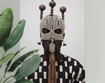May include: A person wearing a black and white patterned shirt and a mask made of woven material with a wooden crown. The mask has a geometric design and is attached to a wooden stick.