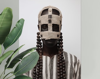 May include: A person is depicted wearing a cream-coloured beaded mask with black detailing, covering the eyes and forehead. Long strands of dark wooden beads hang from the mask. The person is wearing a striped black and white shirt.