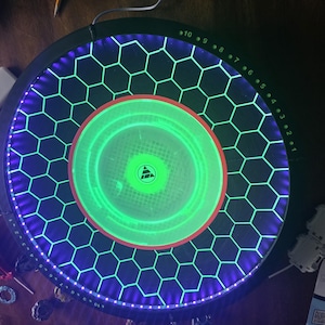 May include: A circular black game arena featuring a glowing green center and a honeycomb pattern illuminated by purple LED lights. The outer rim displays numbered markings from 1 to 10 in green, and a triangular logo is centered in the middle.