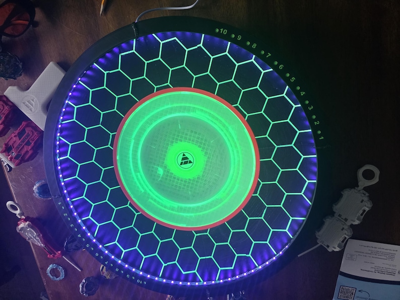 May include: A circular black game arena featuring a glowing green center and a honeycomb pattern illuminated by purple LED lights. The outer rim displays numbered markings from 1 to 10 in green, and a triangular logo is centered in the middle.