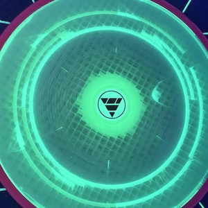 May include: A circular disc with a vibrant neon green glowing center featuring a black triangular logo. The disc has a dark red outer rim and is set against a dark background with a white hexagonal grid pattern that glows under UV light.