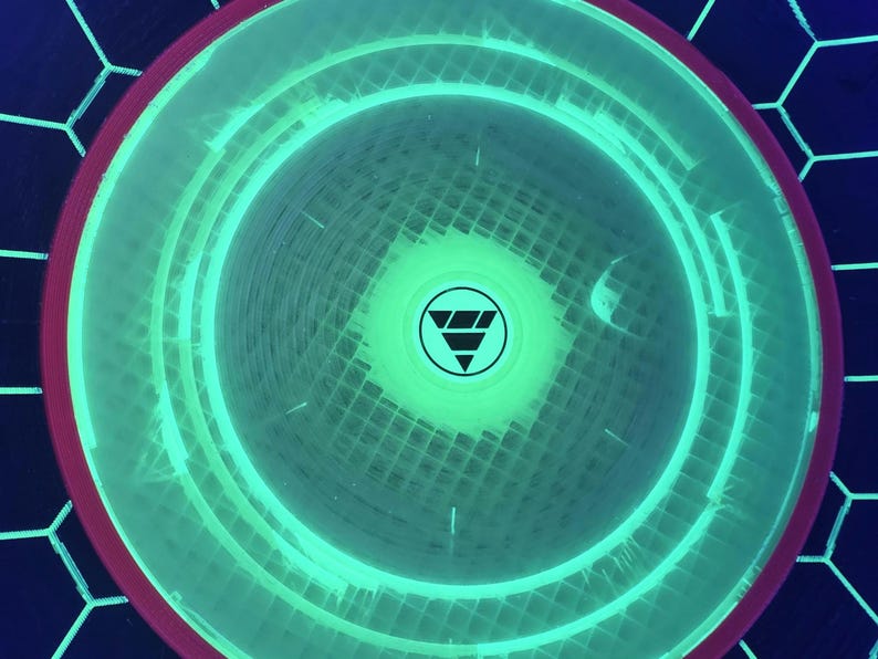 May include: A circular disc with a vibrant neon green glowing center featuring a black triangular logo. The disc has a dark red outer rim and is set against a dark background with a white hexagonal grid pattern that glows under UV light.
