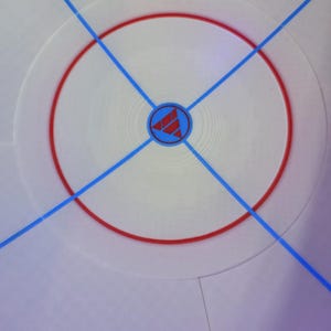 May include: A white circular surface with a red ring and blue lines forming an X. A blue and red logo is in the center. The design is reminiscent of a sports field or game board.