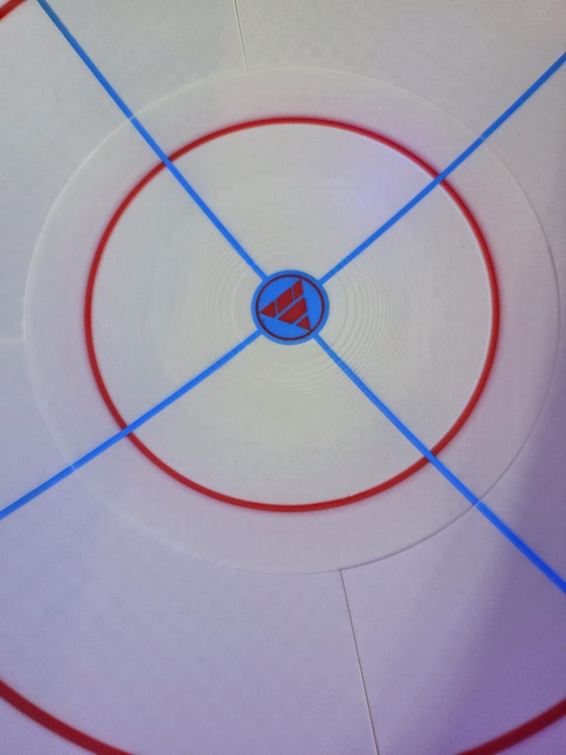May include: A white circular surface with a red ring and blue lines forming an X. A blue and red logo is in the center. The design is reminiscent of a sports field or game board.