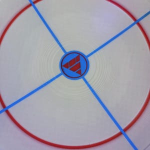 May include: Circular white game board with a red ring and blue lines radiating from the center. A blue circle in the center contains a red geometric design. The board has a textured surface.