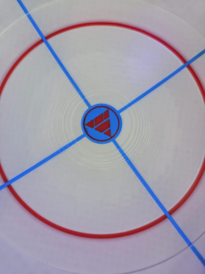 May include: Circular white game board with a red ring and blue lines radiating from the center. A blue circle in the center contains a red geometric design. The board has a textured surface.