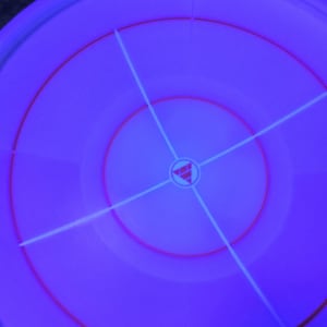 May include: A circular, purple object with concentric rings and a central design. Red and white lines intersect across the surface, creating a target-like pattern. The object has a glowing, neon-like appearance.