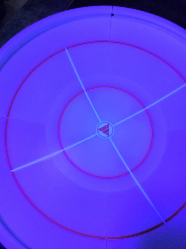 May include: A circular, purple object with concentric rings and a central design. Red and white lines intersect across the surface, creating a target-like pattern. The object has a glowing, neon-like appearance.