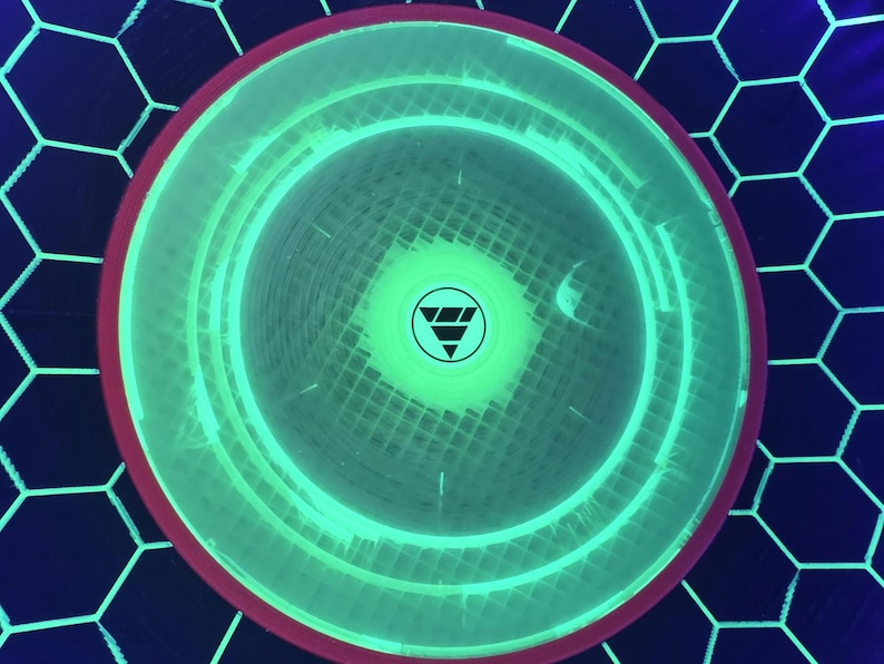 May include: A circular disc with a vibrant neon green glowing center and a dark red outer rim. The surface features a grid pattern and a central circular logo containing a downward-pointing triangle composed of three horizontal bars.