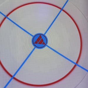 May include: Circular white object with a red ring and blue lines radiating from the center. A blue circle with a red triangle design is in the center. The object has a textured surface.