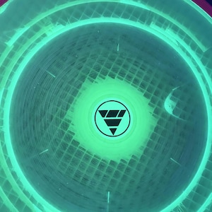 May include: A circular disc with a vibrant neon green glow and a translucent grid pattern. The center features a black circular emblem containing a stylized triangle design composed of three horizontal bars stacked vertically within a circle.