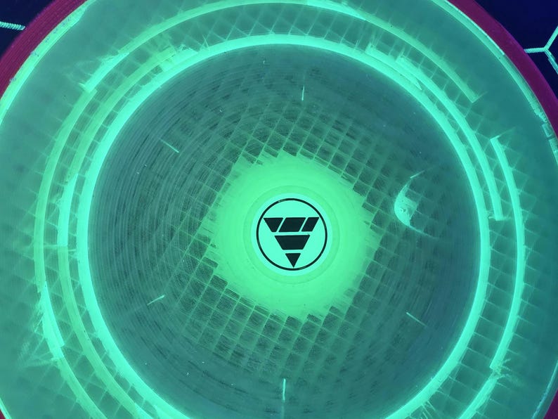 May include: A circular disc with a vibrant neon green glow and a translucent grid pattern. The center features a black circular emblem containing a stylized triangle design composed of three horizontal bars stacked vertically within a circle.