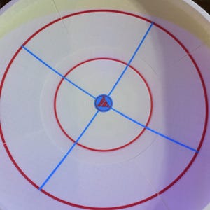 May include: A white plastic arena with red and blue markings. The arena has a circular design with a central emblem and radiating blue lines. The outer rim is white with a slight yellow hue.