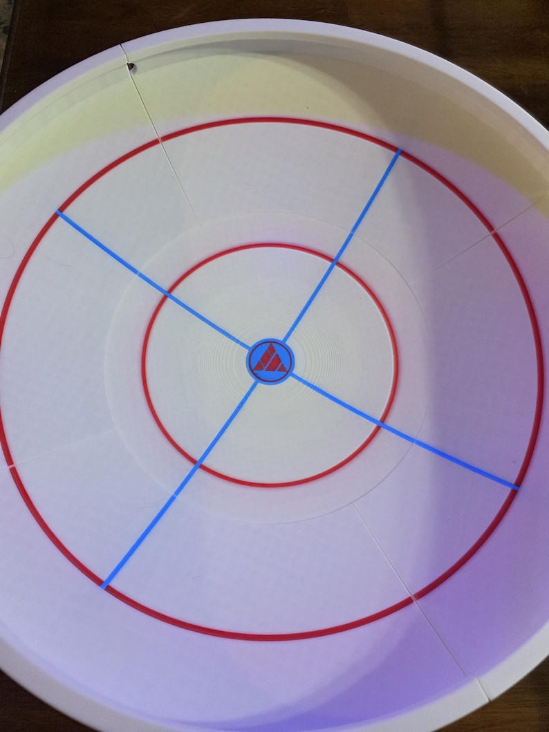 May include: A white plastic arena with red and blue markings. The arena has a circular design with a central emblem and radiating blue lines. The outer rim is white with a slight yellow hue.