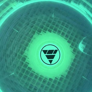 May include: A circular object with a vibrant neon green glow, featuring a grid-like texture across its surface. In the center, there is a black circular emblem containing a stylized downward-pointing triangle composed of three stacked horizontal segments.