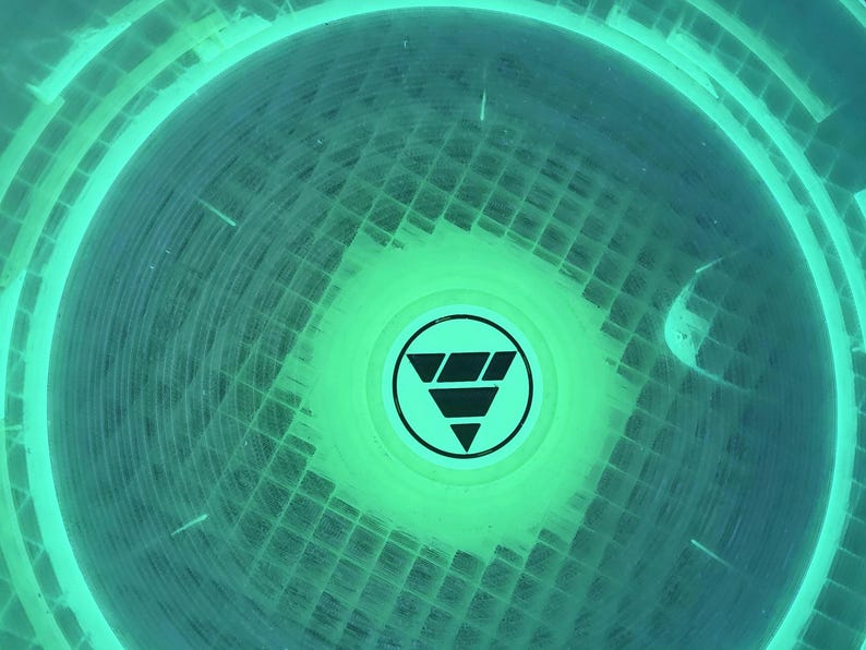 May include: A circular object with a vibrant neon green glow, featuring a grid-like texture across its surface. In the center, there is a black circular emblem containing a stylized downward-pointing triangle composed of three stacked horizontal segments.