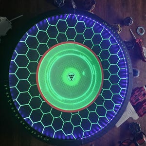 May include: A circular Beyblade stadium featuring a black surface with a glowing green hexagonal grid pattern. The outer rim is illuminated with purple LED lights and includes numbered markings from 1 to 10. A red ring surrounds the center.