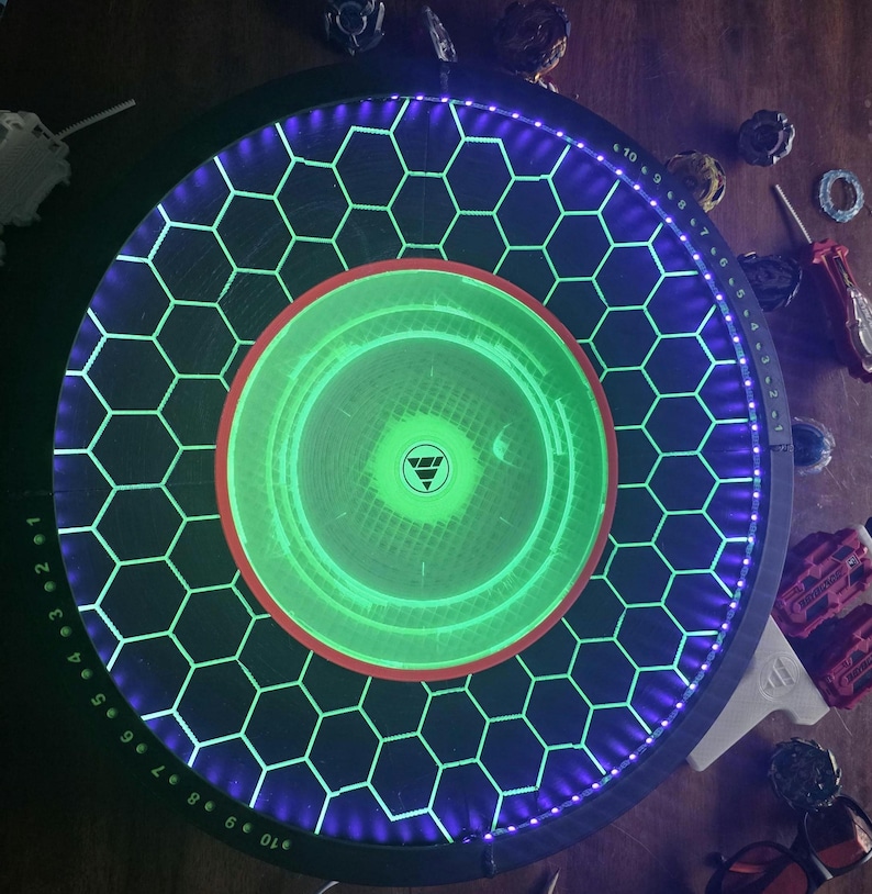 May include: A circular Beyblade stadium featuring a black surface with a glowing green hexagonal grid pattern. The outer rim is illuminated with purple LED lights and includes numbered markings from 1 to 10. A red ring surrounds the center.