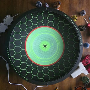 May include: A circular black Beyblade stadium featuring a green hexagonal grid pattern, a central lime green circle with a red border, and numbered scoring markers from 1 to 10 along the outer rim, accompanied by various spinning tops and launchers.