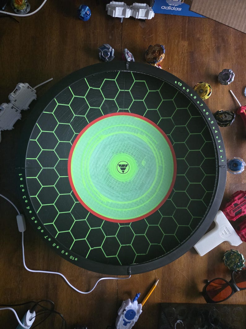 May include: A circular black Beyblade stadium featuring a green hexagonal grid pattern, a central lime green circle with a red border, and numbered scoring markers from 1 to 10 along the outer rim, accompanied by various spinning tops and launchers.