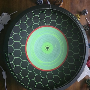 May include: A circular black Beyblade stadium featuring a green hexagonal grid pattern, a central lime green circle with a red border, and a white logo. The outer rim displays numerical markings from 1 to 10 in white text for scoring purposes.