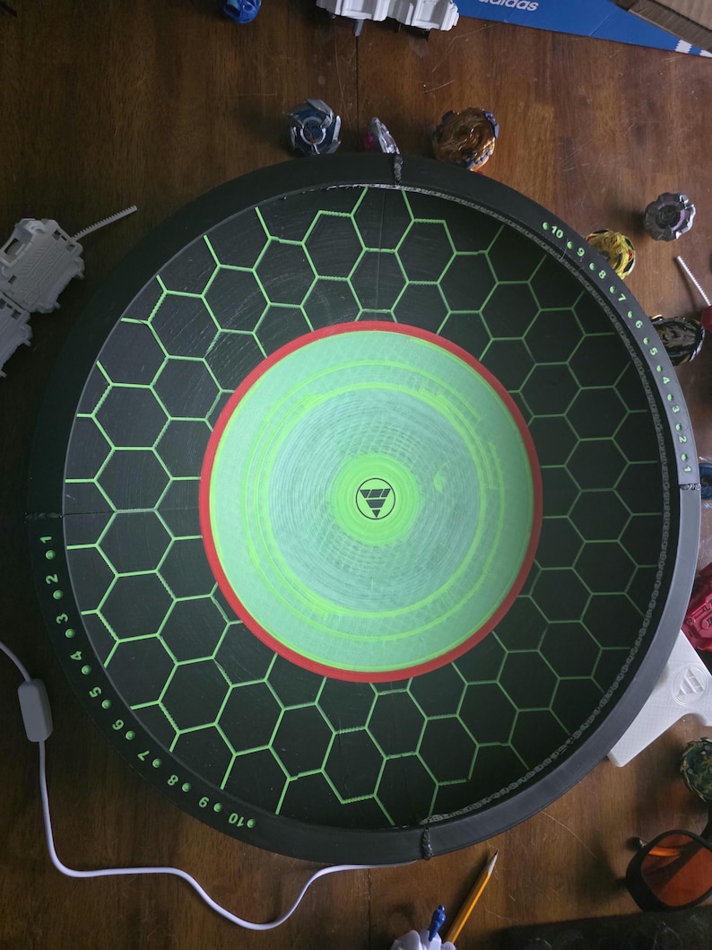 May include: A circular black Beyblade stadium featuring a green hexagonal grid pattern, a central lime green circle with a red border, and a white logo. The outer rim displays numerical markings from 1 to 10 in white text for scoring purposes.