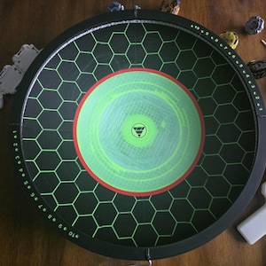 May include: A circular black Beyblade stadium featuring a green hexagonal grid pattern, a central lime green circle with a red border, and numbered markings from 1 to 10 along the inner rim, accompanied by various spinning tops and launchers.