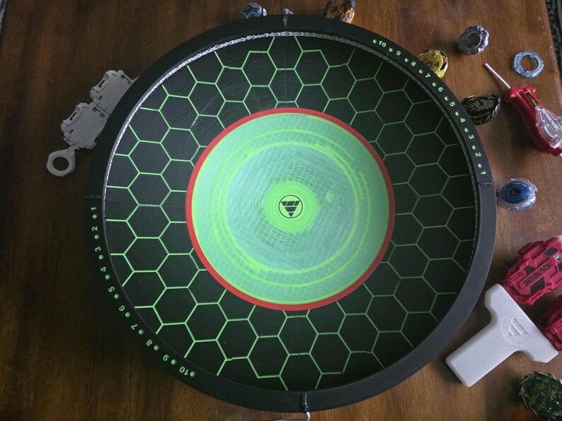 May include: A circular black Beyblade stadium featuring a green hexagonal grid pattern, a central lime green circle with a red border, and numbered markings from 1 to 10 along the inner rim, accompanied by various spinning tops and launchers.