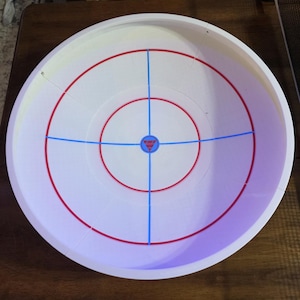 May include: A white plastic circular game arena with red and blue markings. The arena has concentric red circles and blue lines intersecting at the center. A small red and blue emblem is at the center of the arena.