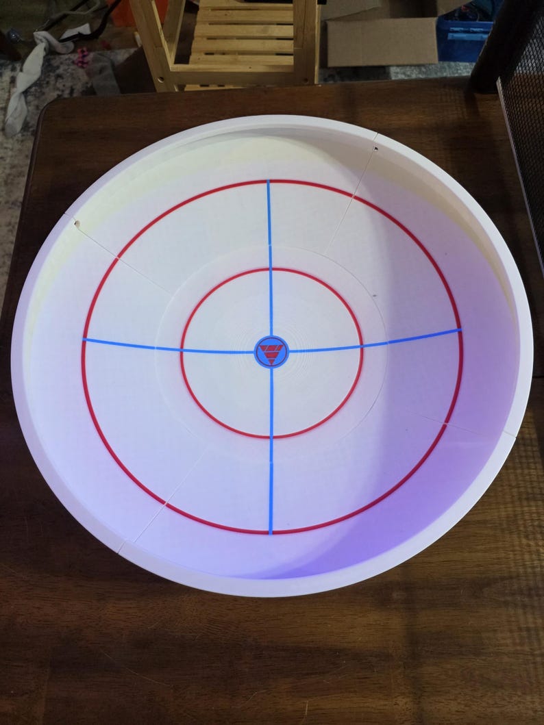 May include: A white plastic circular game arena with red and blue markings. The arena has concentric red circles and blue lines intersecting at the center. A small red and blue emblem is at the center of the arena.
