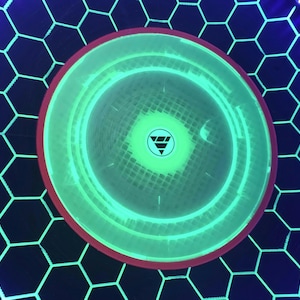 May include: A circular light-up display featuring a bright green glowing center with a small triangular logo. The surrounding area has a black hexagonal grid pattern illuminated by thin green lines, framed by a thin red ring and a blue LED border.