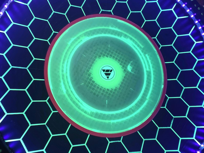 May include: A circular light-up display featuring a bright green glowing center with a small triangular logo. The surrounding area has a black hexagonal grid pattern illuminated by thin green lines, framed by a thin red ring and a blue LED border.