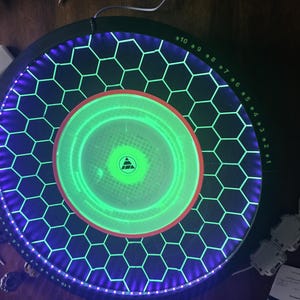 May include: A circular black game arena featuring a central glowing green disc with a triangular logo. The surface is patterned with a black honeycomb grid illuminated by purple LED lights along the perimeter and numbered markers from 1 to 10.