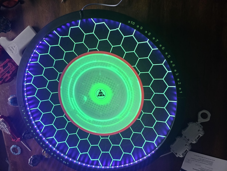 May include: A circular black game arena featuring a central glowing green disc with a triangular logo. The surface is patterned with a black honeycomb grid illuminated by purple LED lights along the perimeter and numbered markers from 1 to 10.