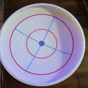 May include: A white plastic game board with red and blue markings. The board has a circular design with concentric red rings and blue lines dividing the surface into quadrants. A small logo is in the center.