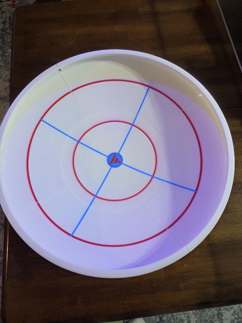 May include: A white plastic game board with red and blue markings. The board has a circular design with concentric red rings and blue lines dividing the surface into quadrants. A small logo is in the center.