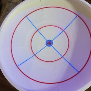 May include: A white circular game board with red and blue markings. The board features concentric red circles, blue lines crossing the center, and a central emblem. The board is designed for a tabletop game.