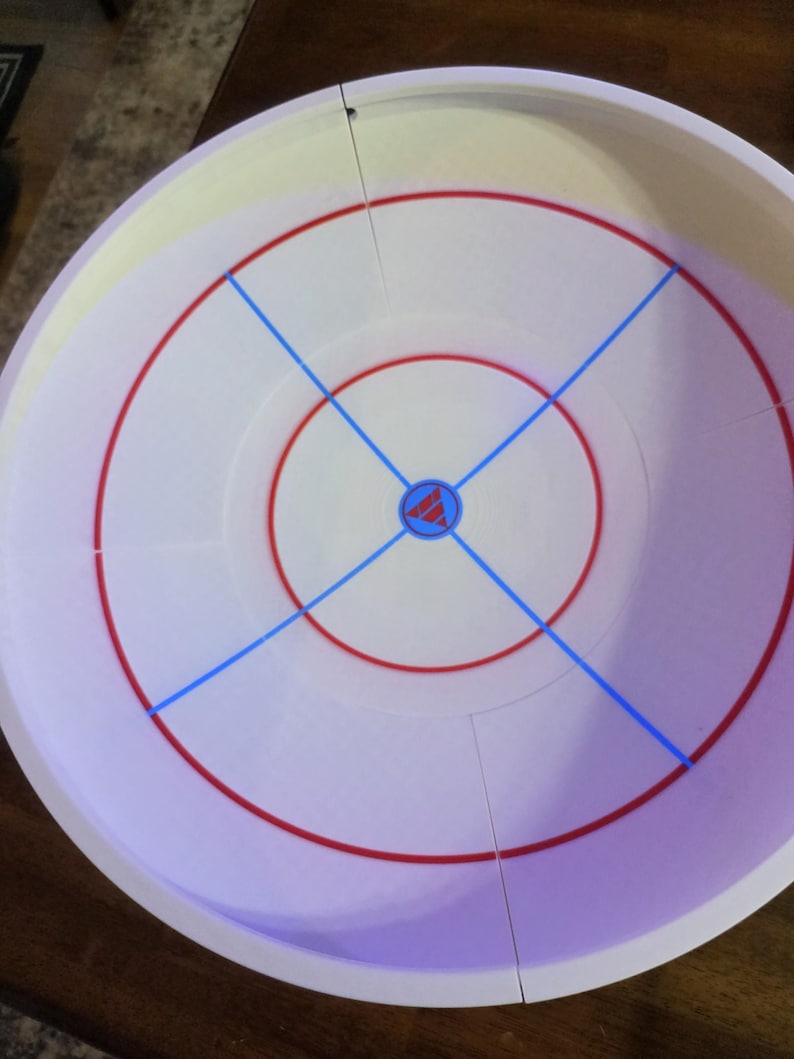 May include: A white circular game board with red and blue markings. The board features concentric red circles, blue lines crossing the center, and a central emblem. The board is designed for a tabletop game.