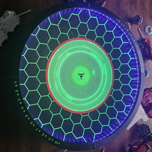May include: A circular Beyblade stadium featuring a black base with a glowing green hexagonal grid pattern, a bright green center circle, and a purple LED-lit outer rim with numbered markings from 1 to 10 printed in white along the edge.