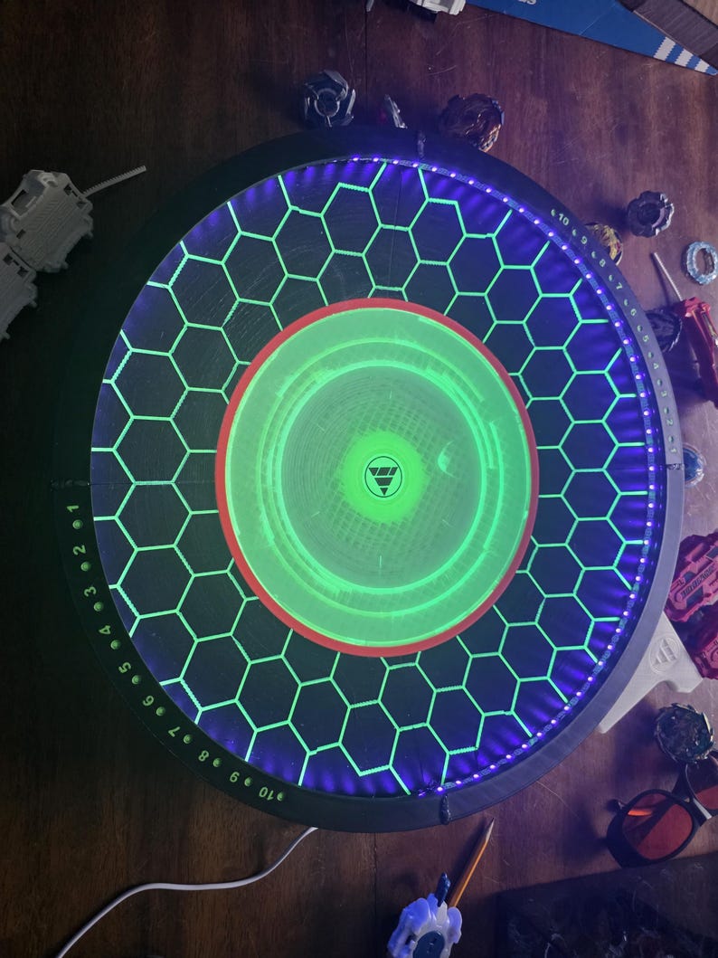 May include: A circular Beyblade stadium featuring a black base with a glowing green hexagonal grid pattern, a bright green center circle, and a purple LED-lit outer rim with numbered markings from 1 to 10 printed in white along the edge.