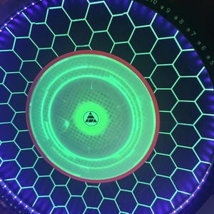 May include: A circular game board featuring a black surface with a glowing green honeycomb grid pattern. The center has a bright green circular light with a small black triangle logo. A purple LED ring borders the edge with numbered markers.