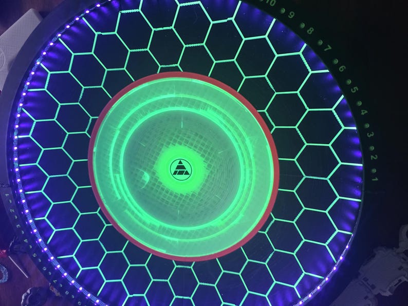 May include: A circular game board featuring a black surface with a glowing green honeycomb grid pattern. The center has a bright green circular light with a small black triangle logo. A purple LED ring borders the edge with numbered markers.