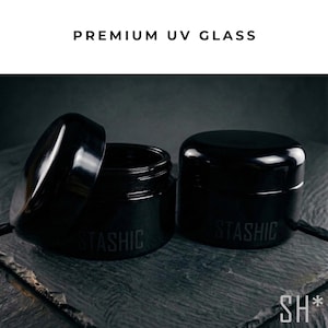 May include: Two black UV glass jars, one with the lid open, displaying the text "STASHIC" on the side. The jars are on a dark gray surface, with the text "PREMIUM UV GLASS" above them. The jars are designed to protect contents from UV light.