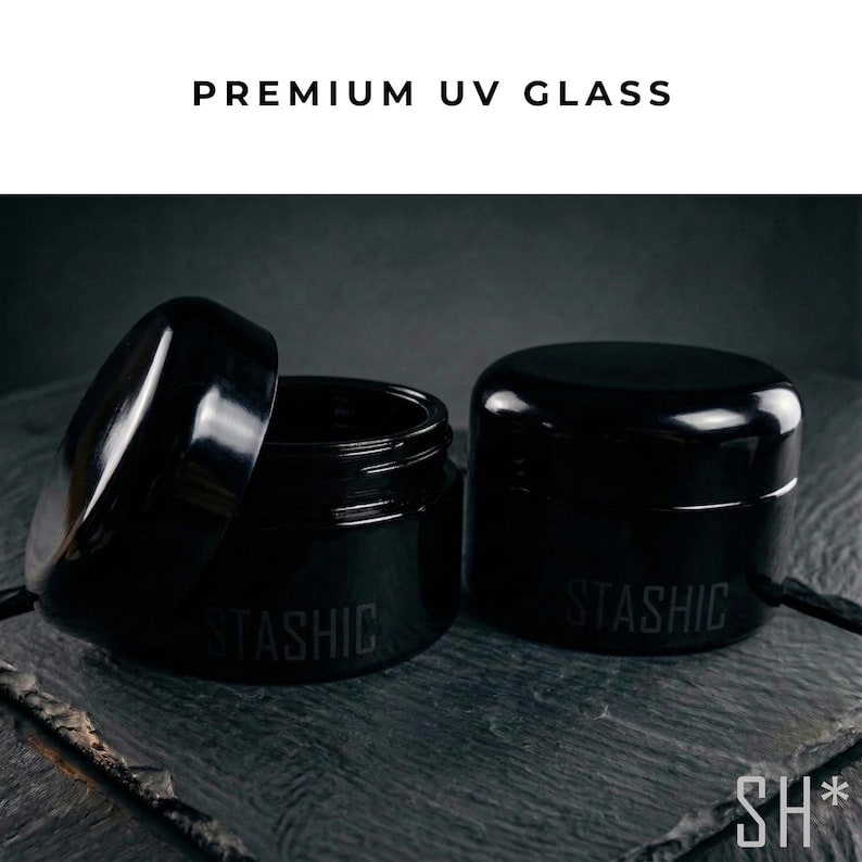 May include: Two black UV glass jars, one with the lid open, displaying the text "STASHIC" on the side. The jars are on a dark gray surface, with the text "PREMIUM UV GLASS" above them. The jars are designed to protect contents from UV light.