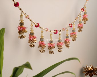 May include: A pink and gold beaded garland with tassels and flower accents. The garland is decorated with small pink and white pom-poms and gold beads. "CRAST INDIA THE HANDICRAFT STORE" is printed on a white background.