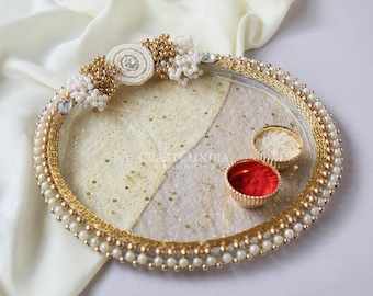 May include: A round, gold-rimmed tray decorated with white pearls and a white floral embellishment. The tray contains two small bowls, one with red powder and the other with white rice.