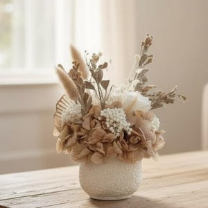 Könnte beinhalten: Dekorative Blumengesteck in einer strukturierten, cremefarbenen Keramikvase. Das Arrangement zeigt Trockenblumen in Braun-, Beige- und Weißtönen, darunter Hortensien, Pampasgras und andere zarte Blüten. Die Vase steht auf einem Holztisch.